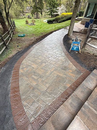 Brick pathway with brown border and steps leading to a yard with trees and a fence.
