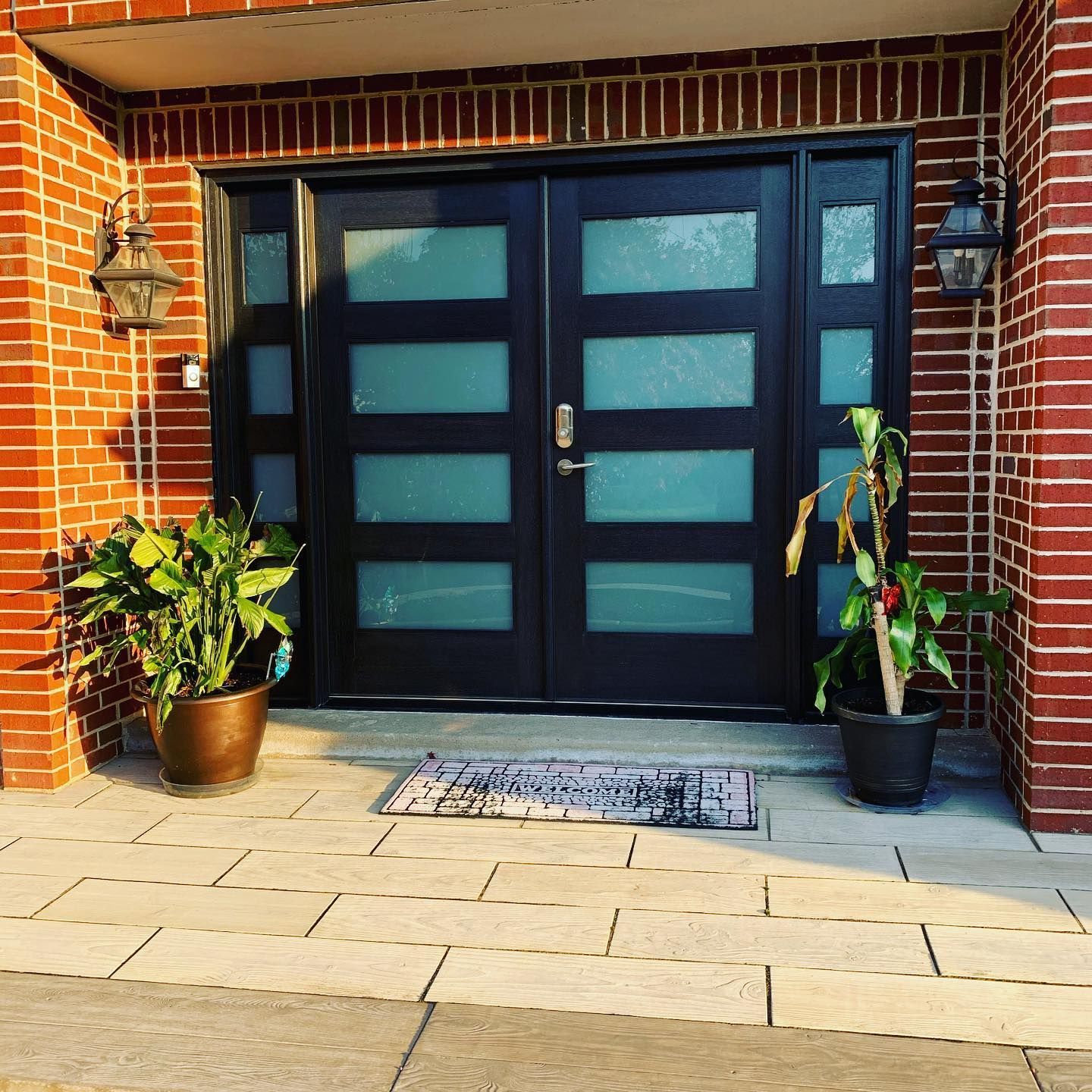 Black double front doors with frosted glass panels, brick facade, potted plants, and welcome mat.