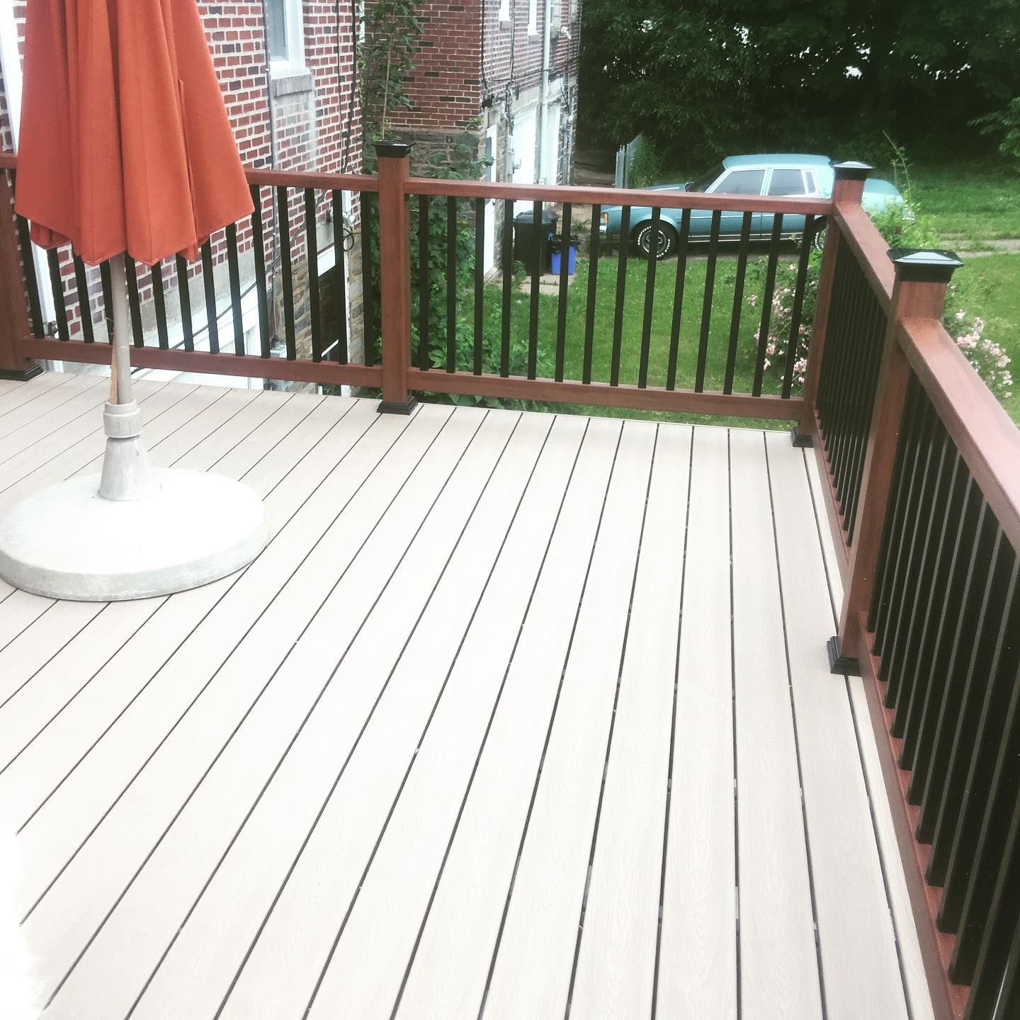 Composite deck with brown railing, black balusters, and orange umbrella. Green lawn and a car are in the background.