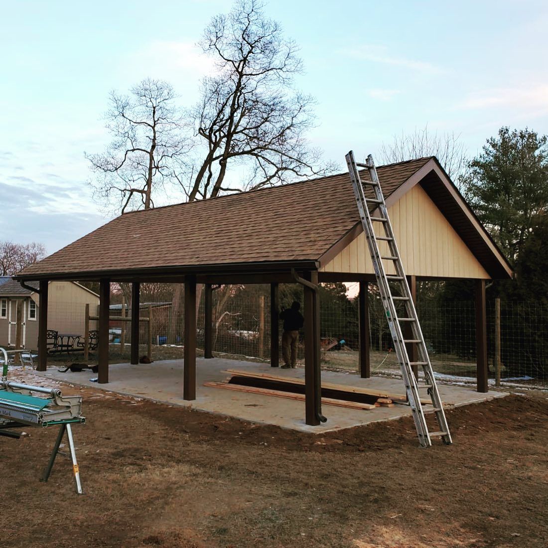 A partially built, brown roofed pavilion on a concrete slab; a ladder leans against it.