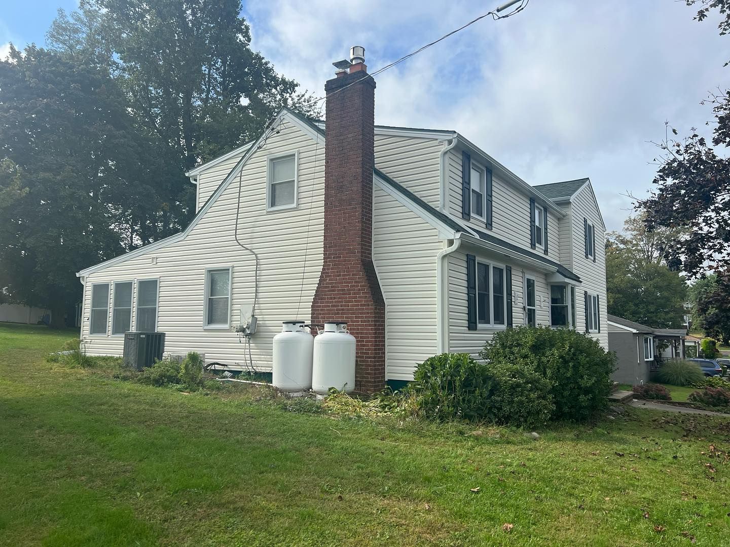 White two-story house with a brick chimney, propane tanks, and green lawn on a cloudy day.
