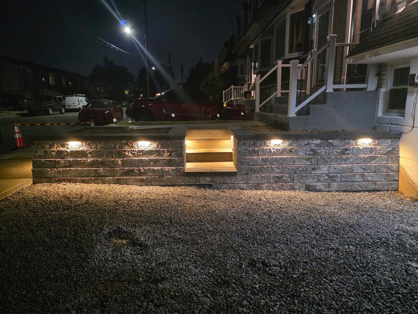 Night view of a stone wall with lit steps, outdoor lights, and gravel. A house is in the background.
