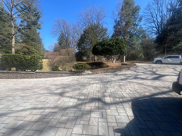 Paver driveway leading to a raised landscape bed with trees under a blue sky.