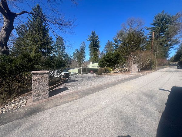 Stone pillars frame the entrance to a driveway. Trees and a light-colored building are visible on a sunny day.