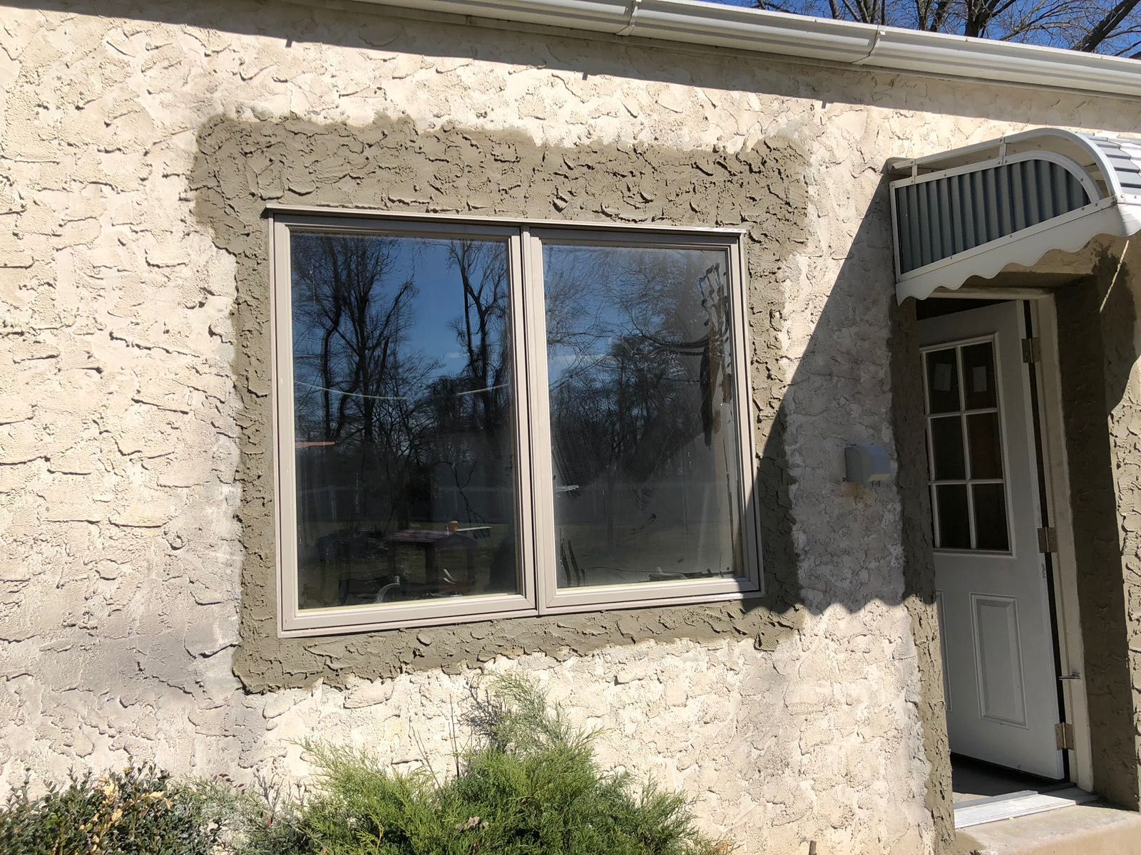Window with freshly applied stucco border on a textured exterior wall, beside a doorway.