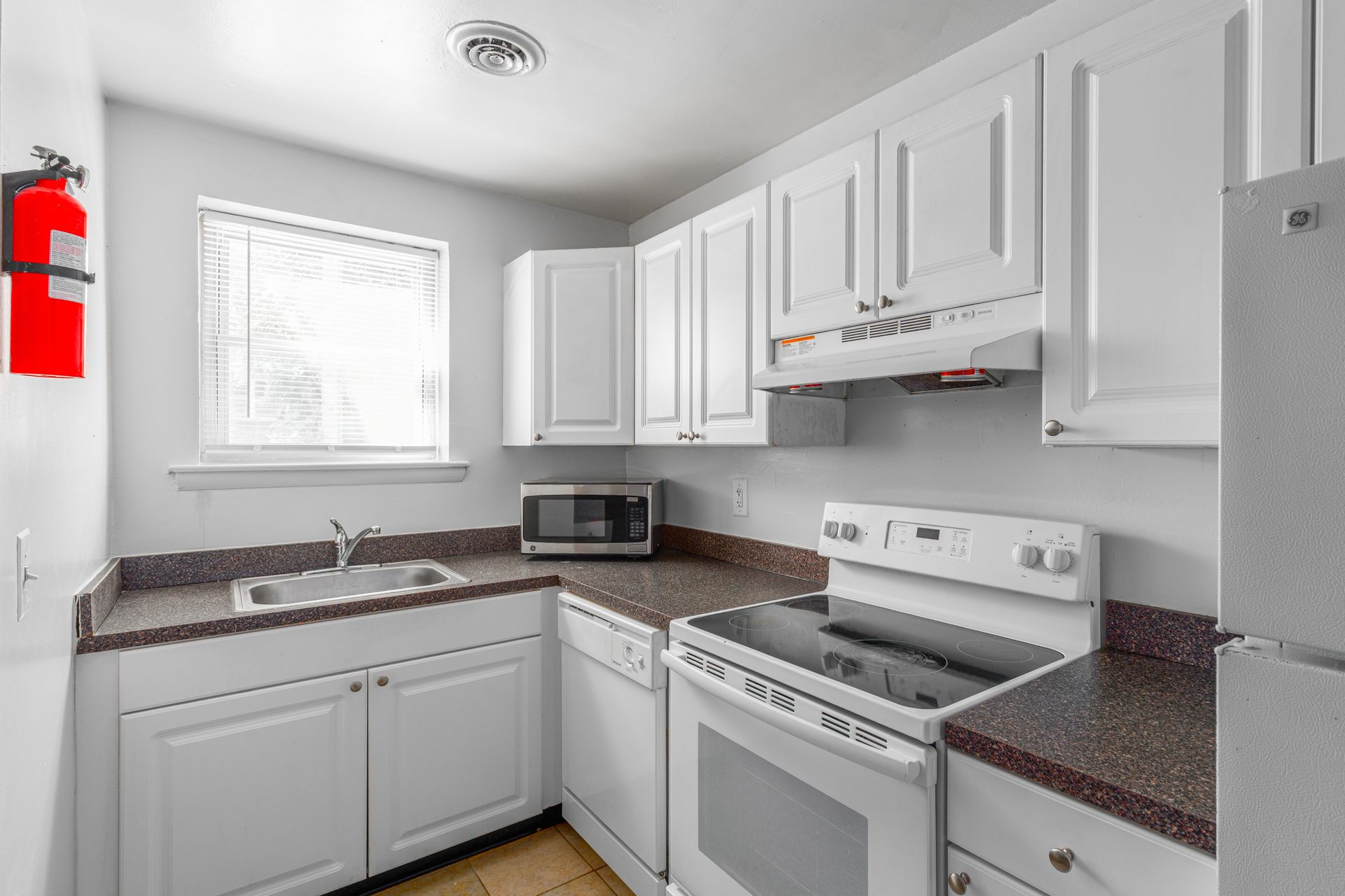 Brookbend Apartments White kitchen with cabinets, stove, microwave, and sink; a fire extinguisher hangs on the wall.