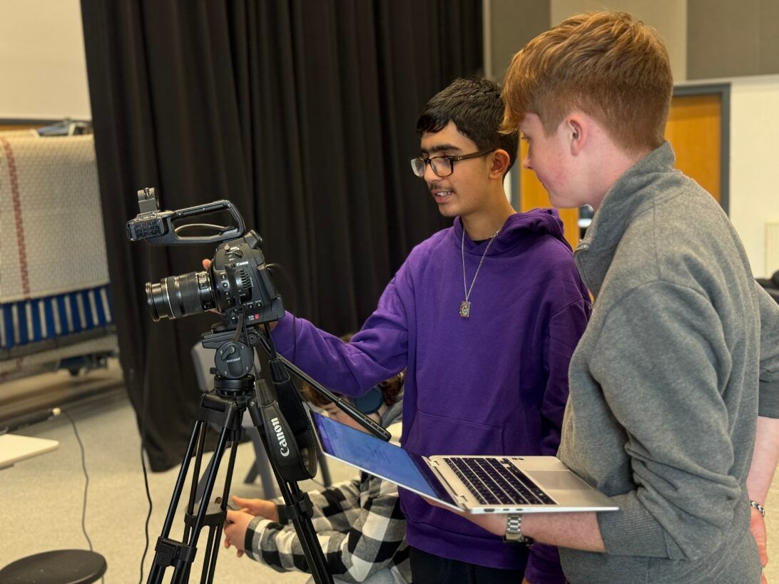 Two young men are looking at a camera while one is holding a laptop.