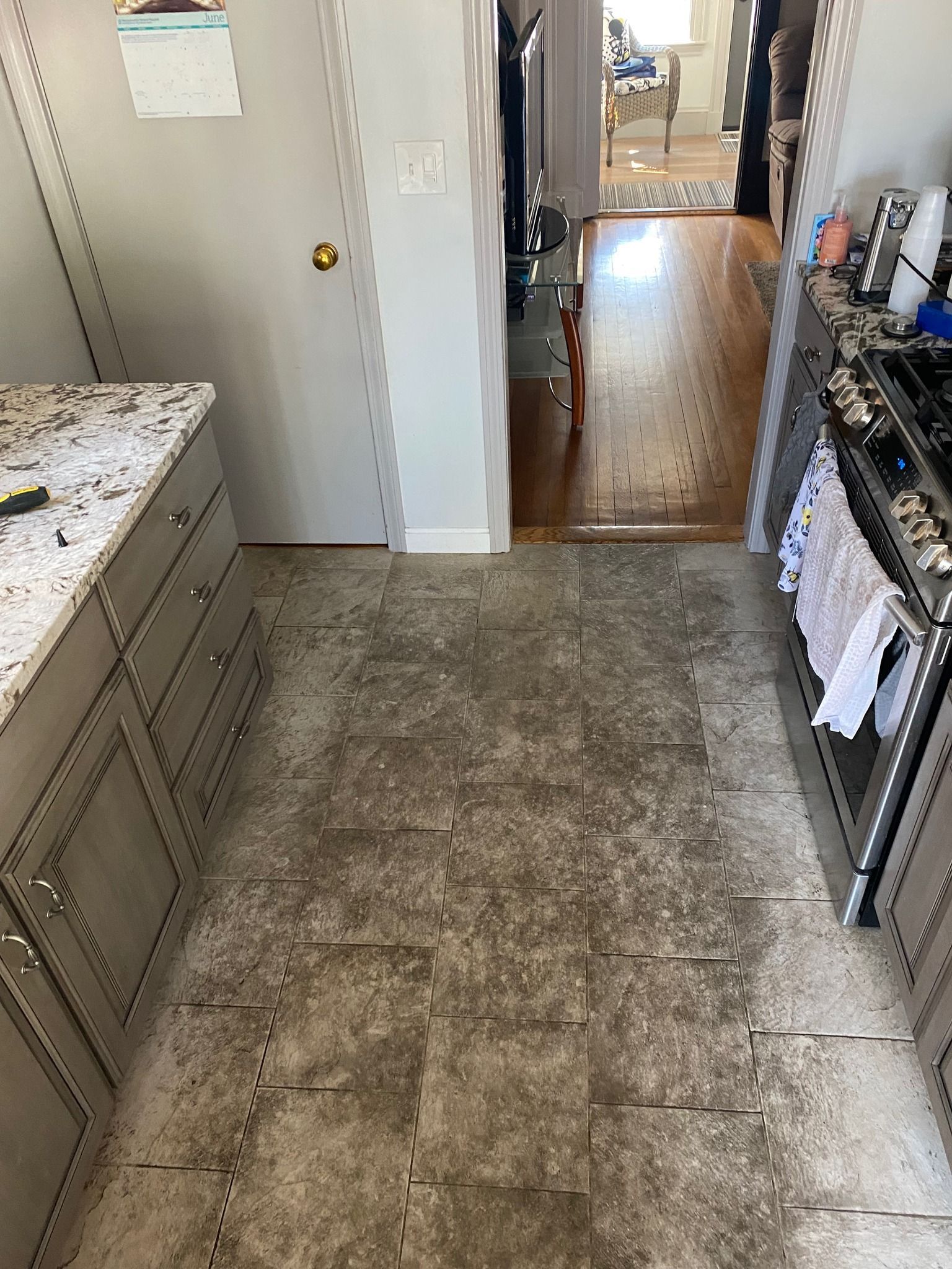 Kitchen with gray cabinets, speckled countertops, and worn floor tiles. Doorway leads to wood floor.