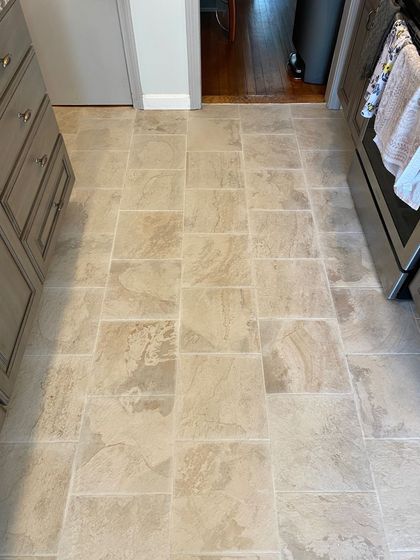 Beige tiled kitchen floor with cabinets and a glimpse into another room with hardwood flooring.