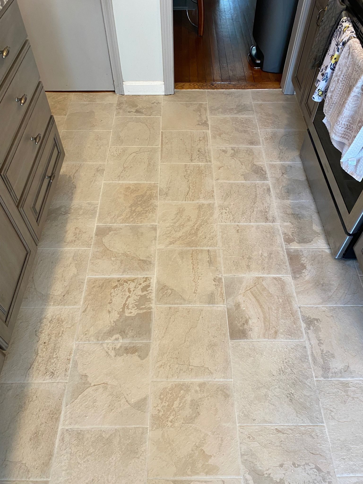 Beige tile floor in a kitchen, with cabinets on the left and a glimpse of another room in the background.