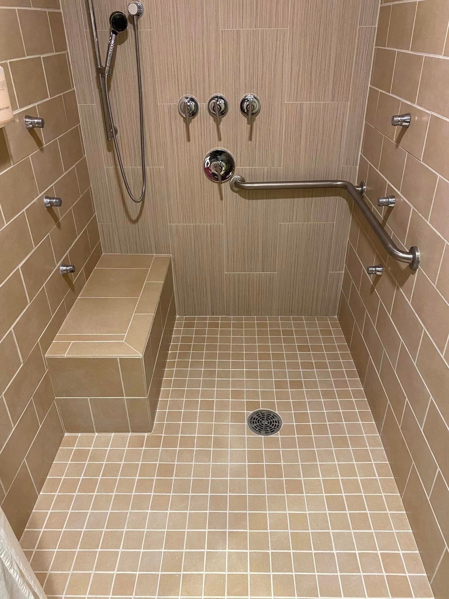 Accessible shower with built-in bench, multiple jets, and grab bar. Tan tile and fixtures.