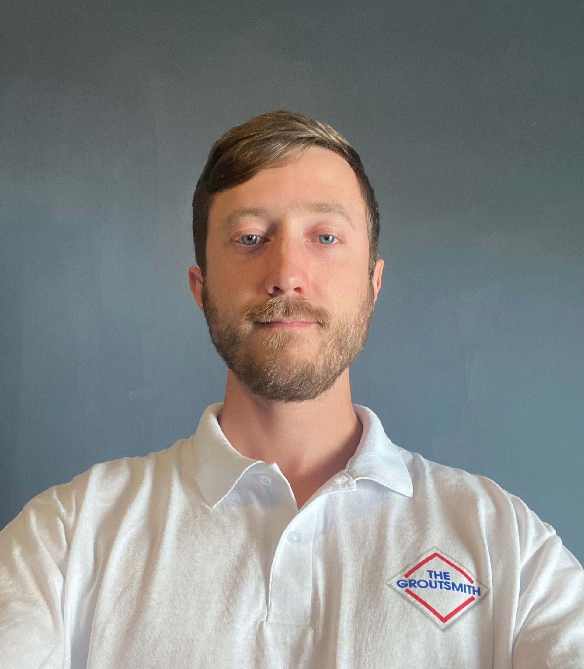 Man with light skin and short brown hair wearing a white polo shirt with an emblem, looking directly at the camera.