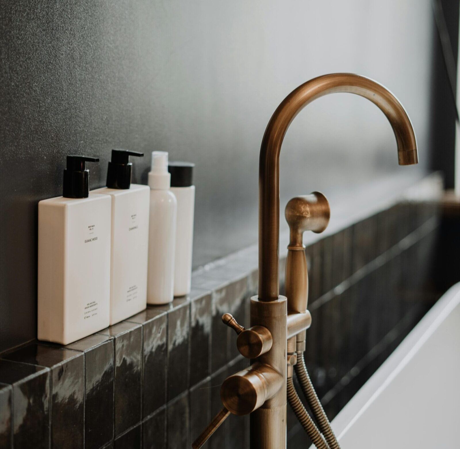 Bathroom with bronze faucet, white bottles, and dark tiles.