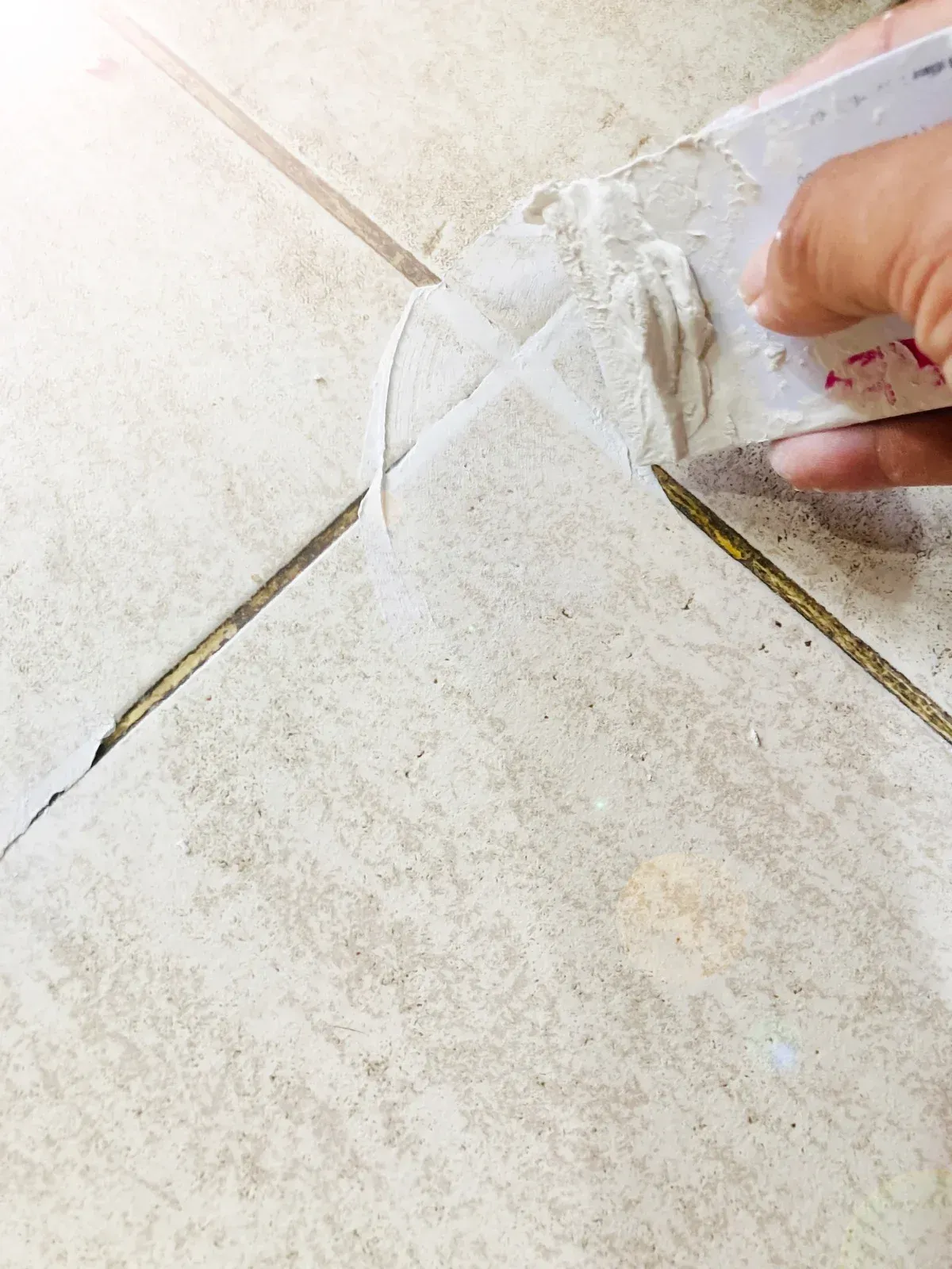 Person using a putty knife to apply grout between beige floor tiles.
