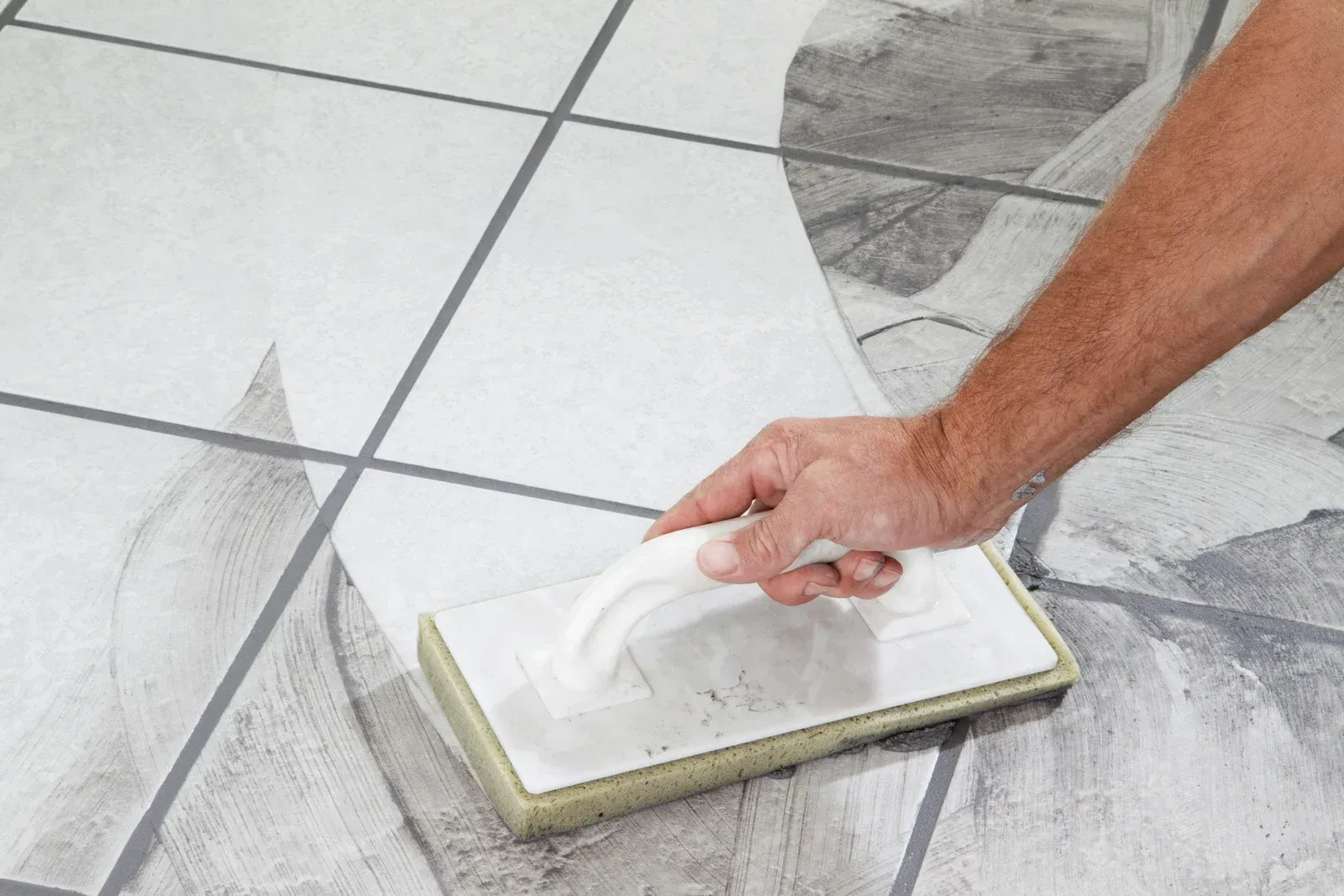 Hand using a grout float to apply white grout between gray floor tiles.