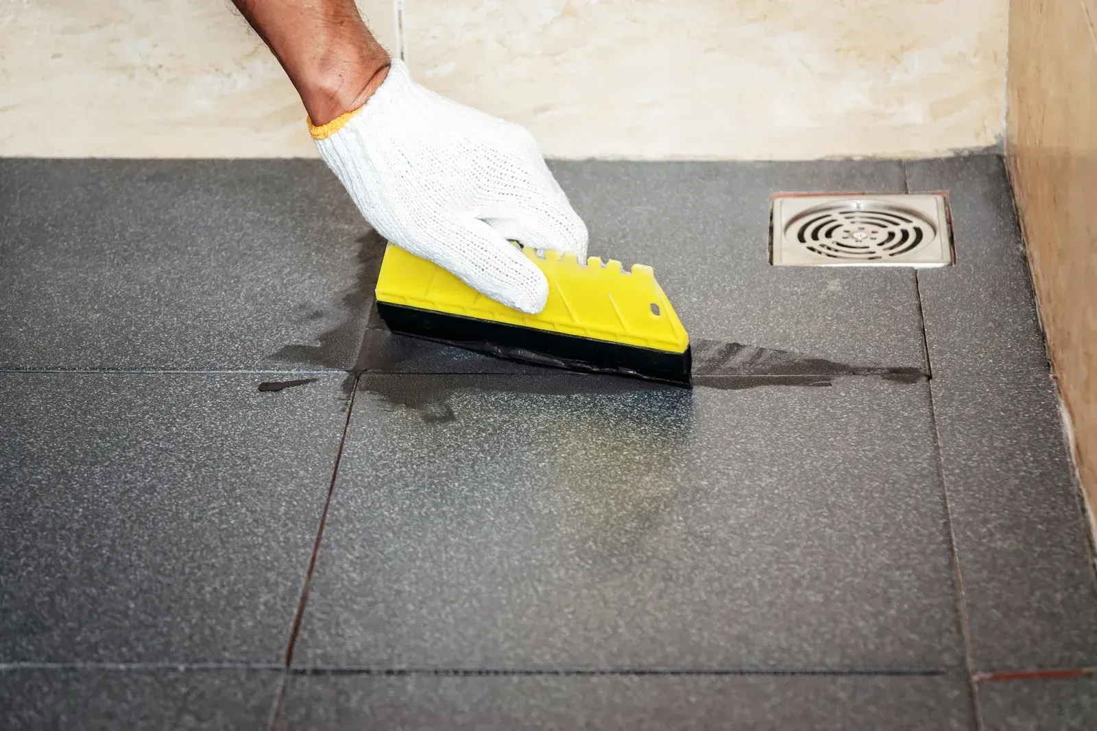 Person in glove applying grout to tile floor with yellow tool near drain.