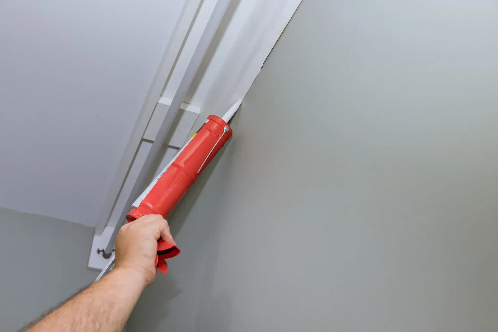 Person applying sealant to molding with a red caulk gun, against a gray wall.