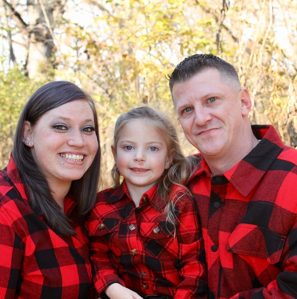 Family of three, wearing matching red and black plaid shirts, smiling outside.
