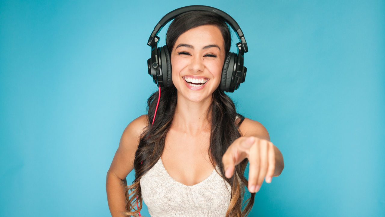 Woman with headphones, smiling and pointing forward, against a blue backdrop.