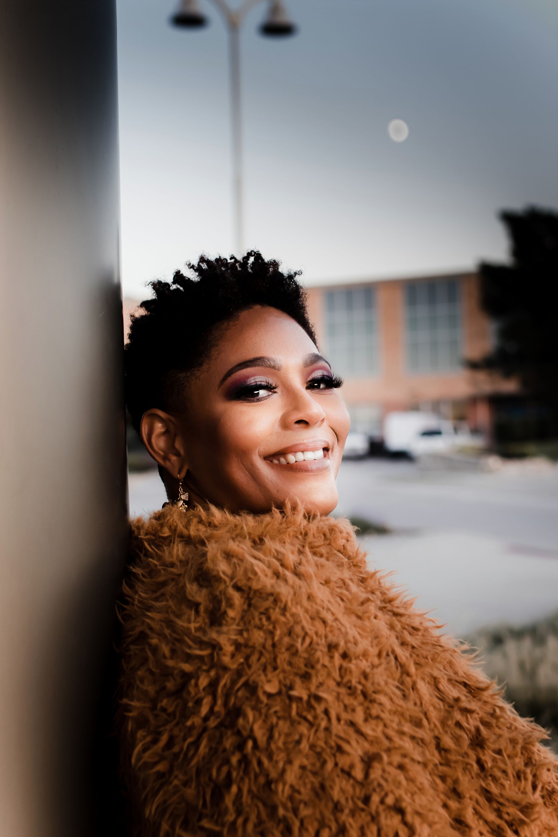 Smiling Black woman leaning against a structure in an outdoor setting, wearing a brown fluffy jacket.