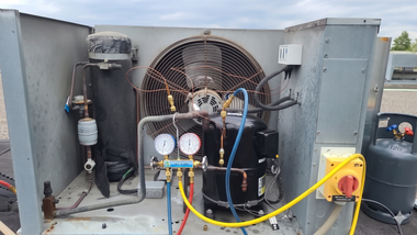 Outdoor HVAC unit with exposed compressor, gauges, hoses, and wiring inside a metal enclosure