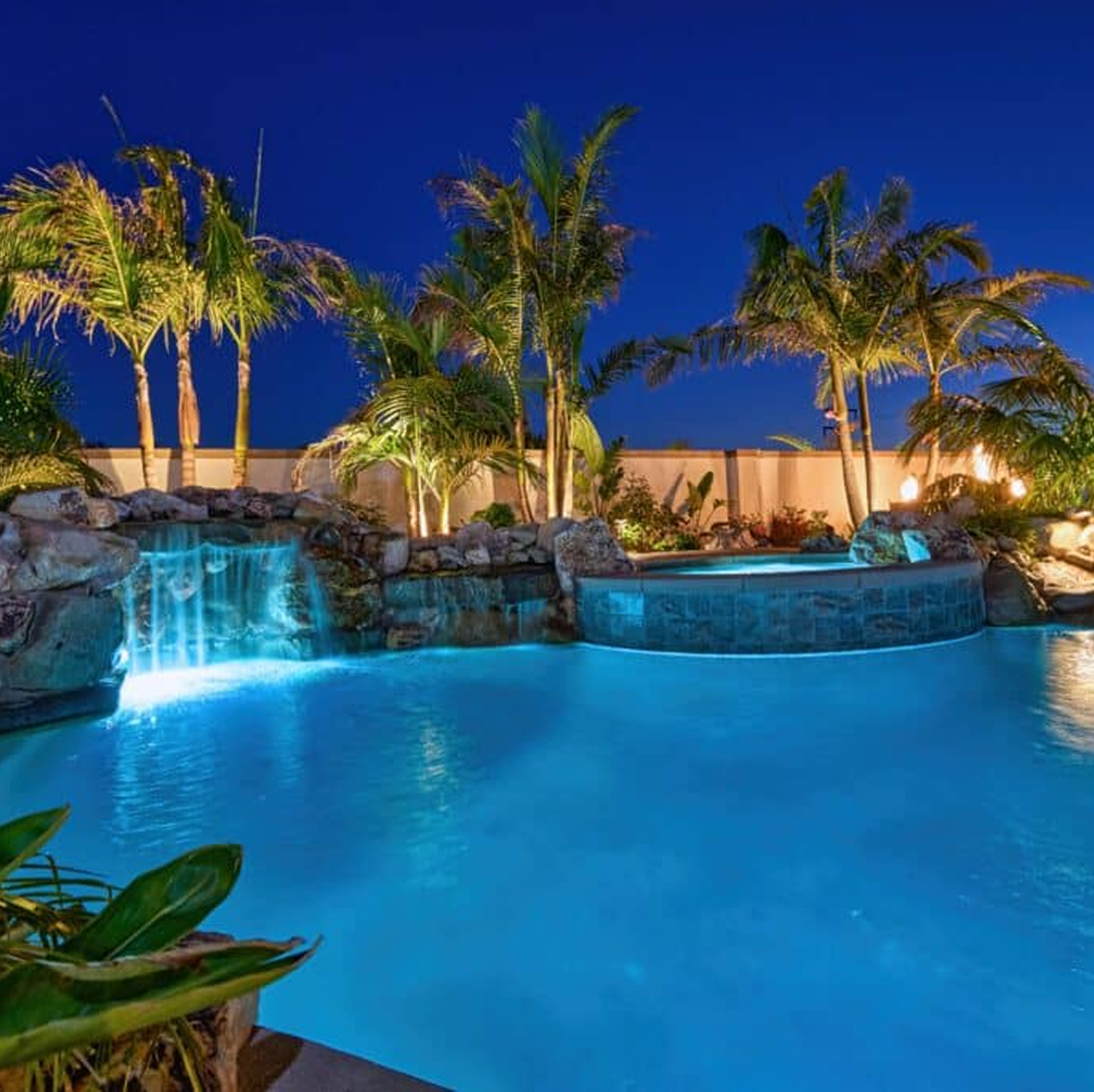 Illuminated pool and waterfall at night with palm trees and a dark blue sky.