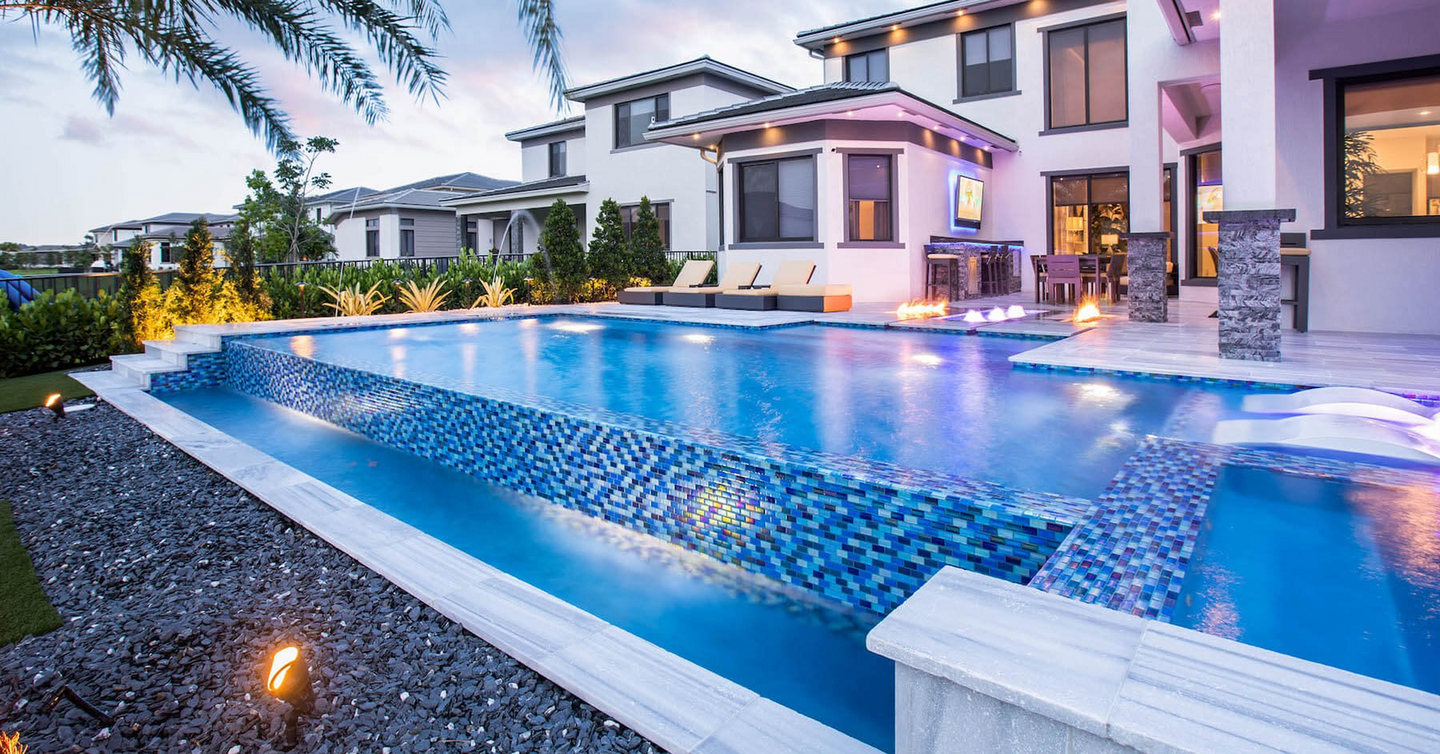 Luxurious backyard pool with blue mosaic tile, lit by underwater lights, next to a modern house.