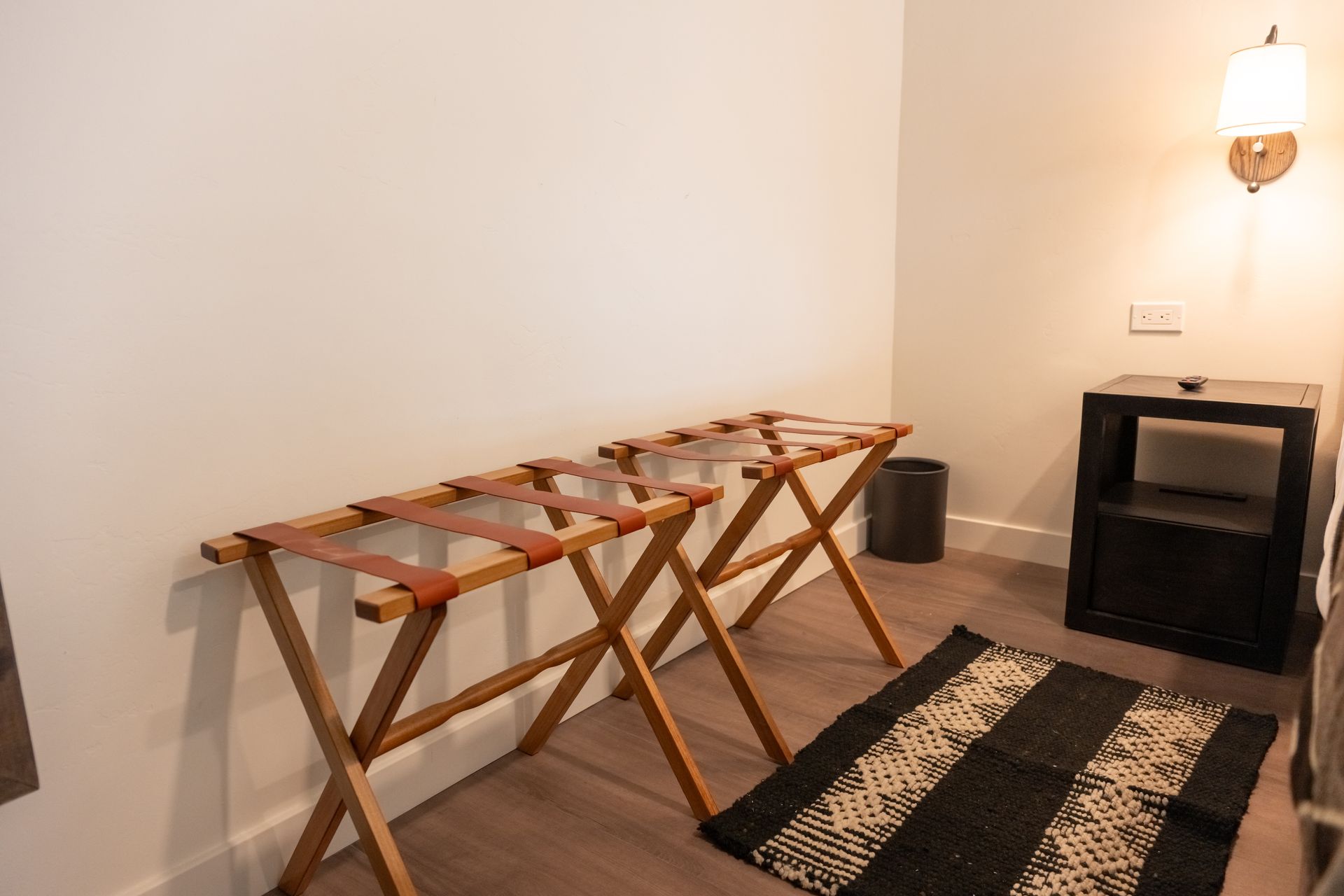 Two wooden luggage racks with leather straps in a room, next to a black nightstand and a patterned rug.