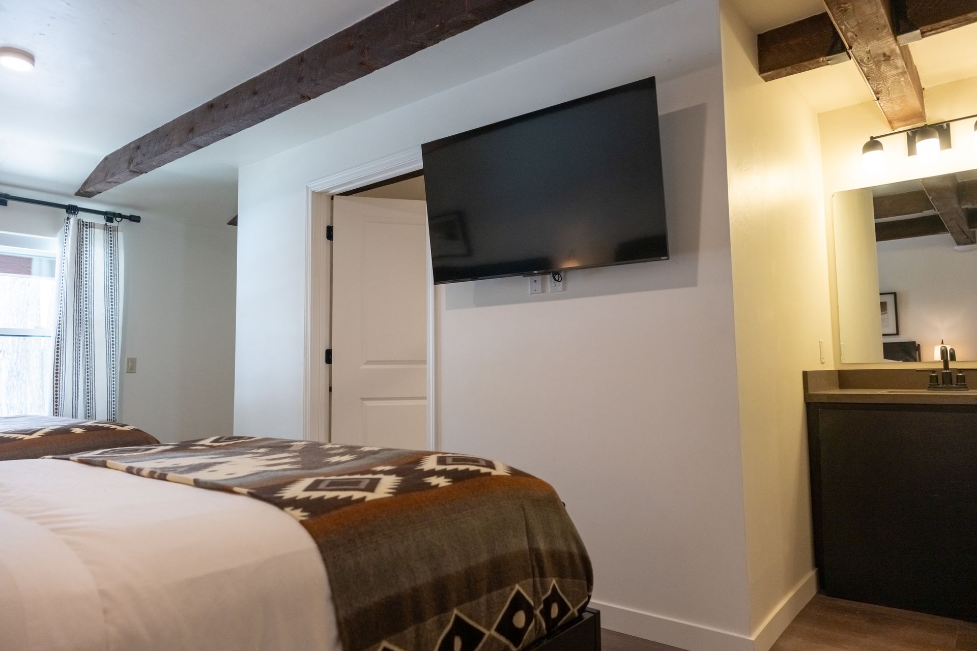 Bedroom with beds, TV, closet, and bathroom with dark cabinets, white walls, and wooden beams.