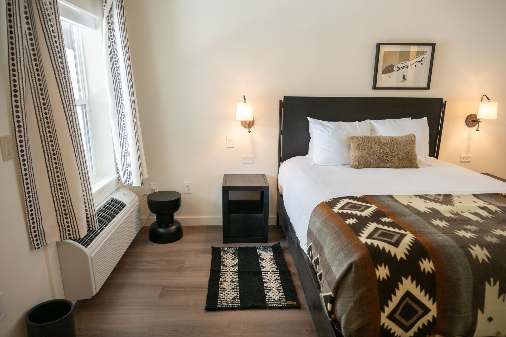 Bedroom with a queen bed, patterned blanket, small rug, window with patterned curtains, and wall sconces.