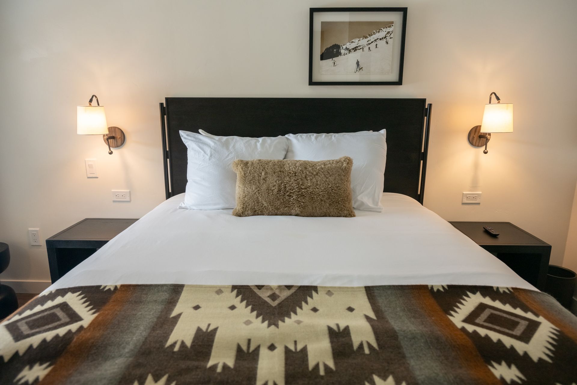 Bedroom with a king-size bed. White bedding, brown patterned blanket, lamps on the wall, and a photo above the headboard.
