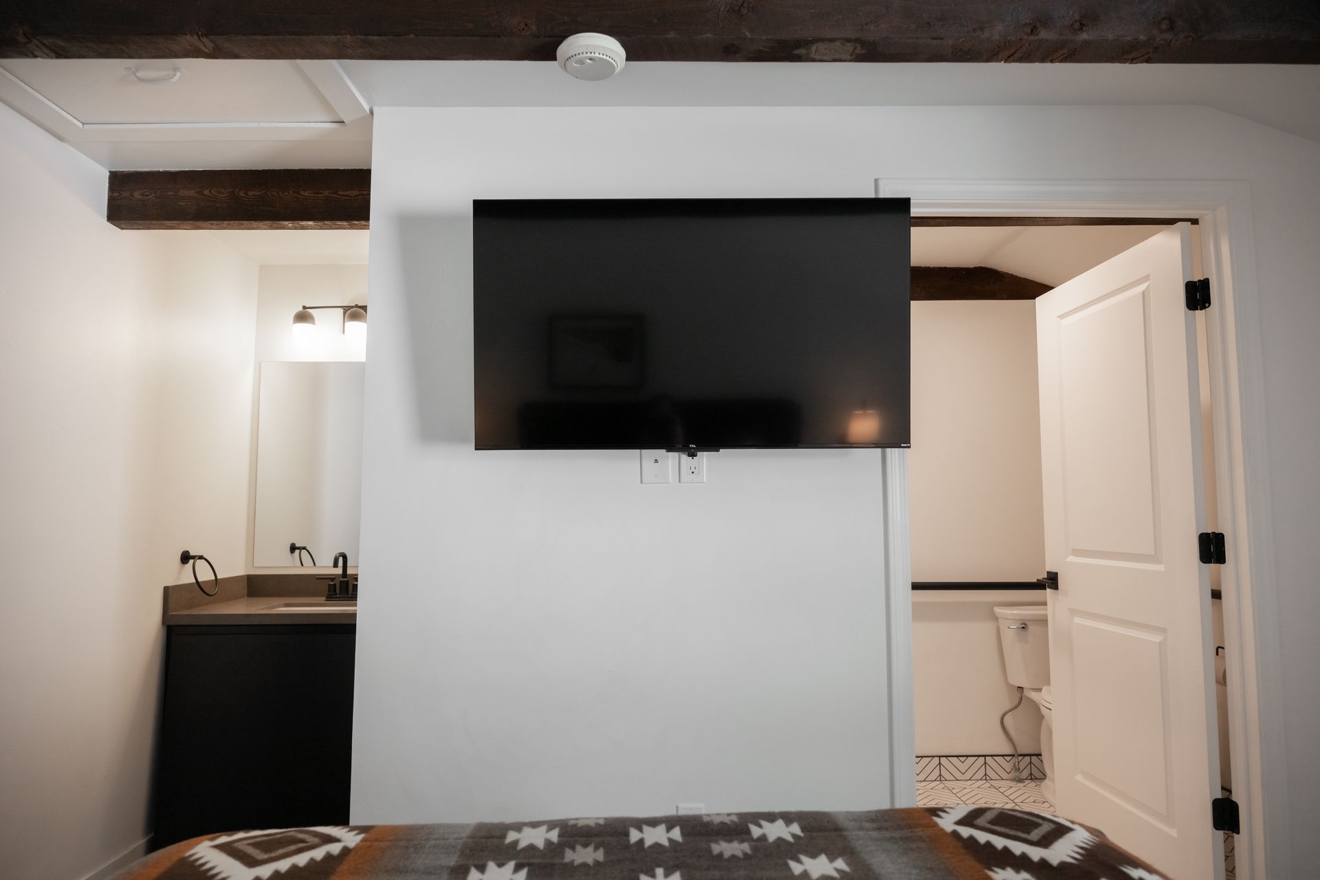 A bedroom with a TV mounted on a white wall, a door to a bathroom, and a dark wood beam.