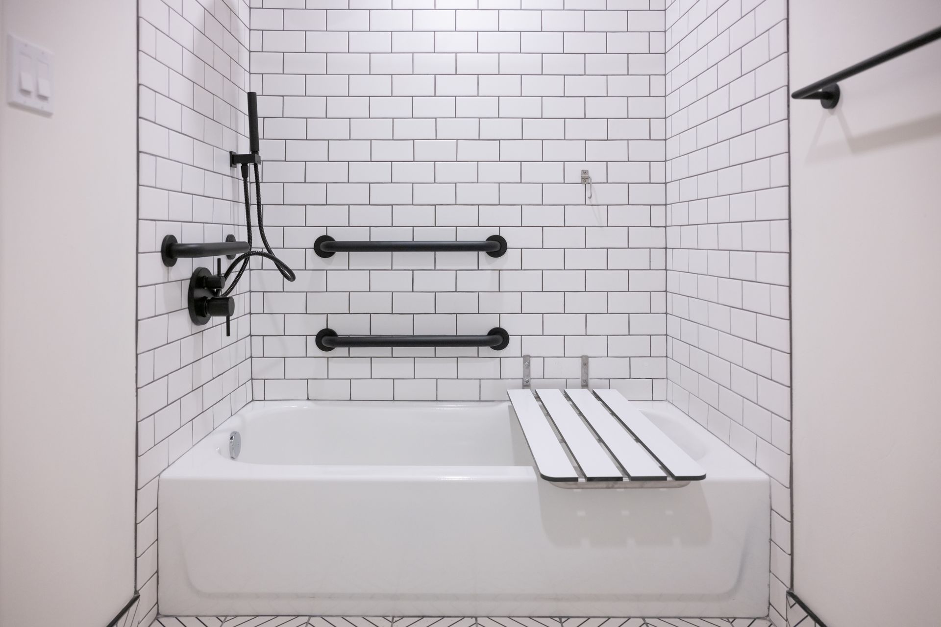 White tiled bathroom with a bathtub, grab bars, and shower fixtures.