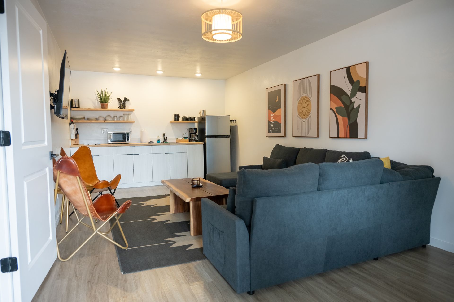 Living room with blue sectional, kitchenette, and art on the wall; wooden floor, and butterfly chair.