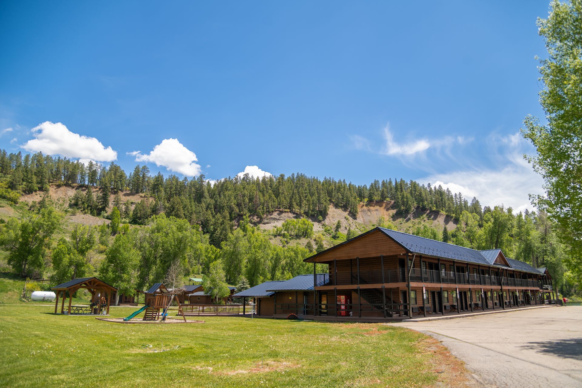 Lodge with a dark roof and two levels, green grass, playground, against a backdrop of trees and hills.