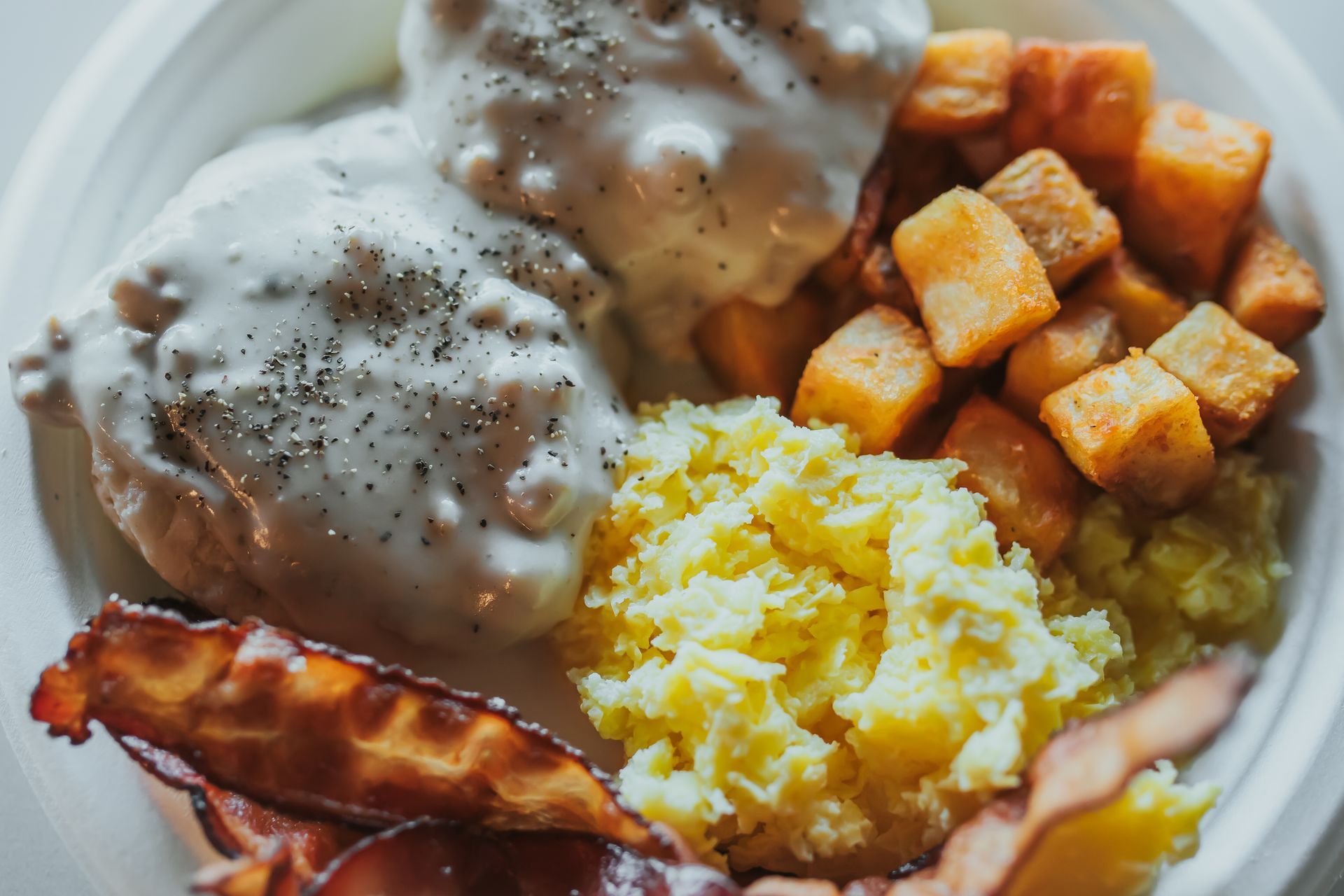 Biscuits and gravy with hash browns, scrambled eggs, and bacon on a plate.