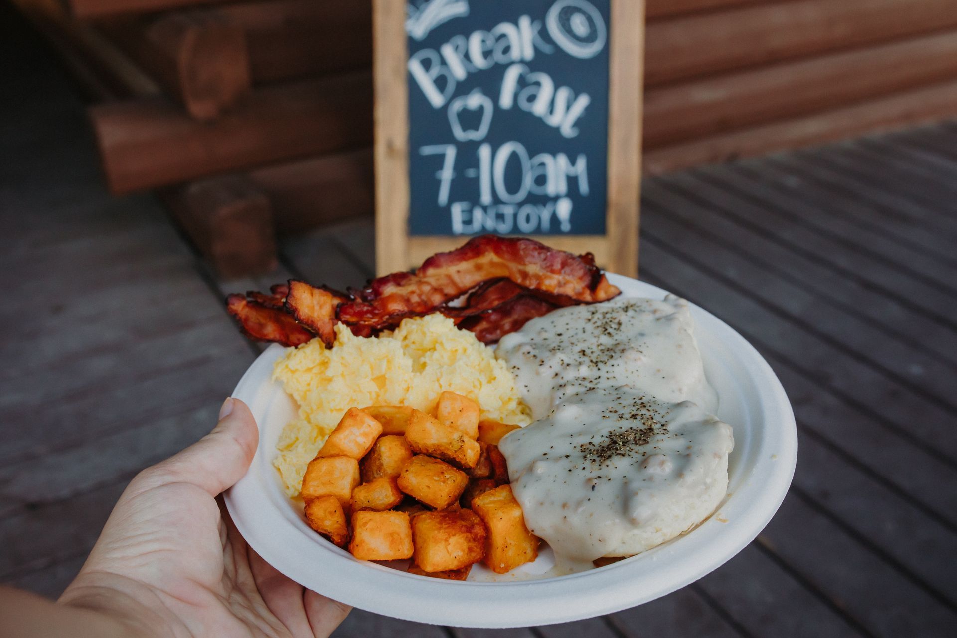 Hand holding plate of breakfast: bacon, scrambled eggs, sweet potatoes, and biscuits covered in gravy.