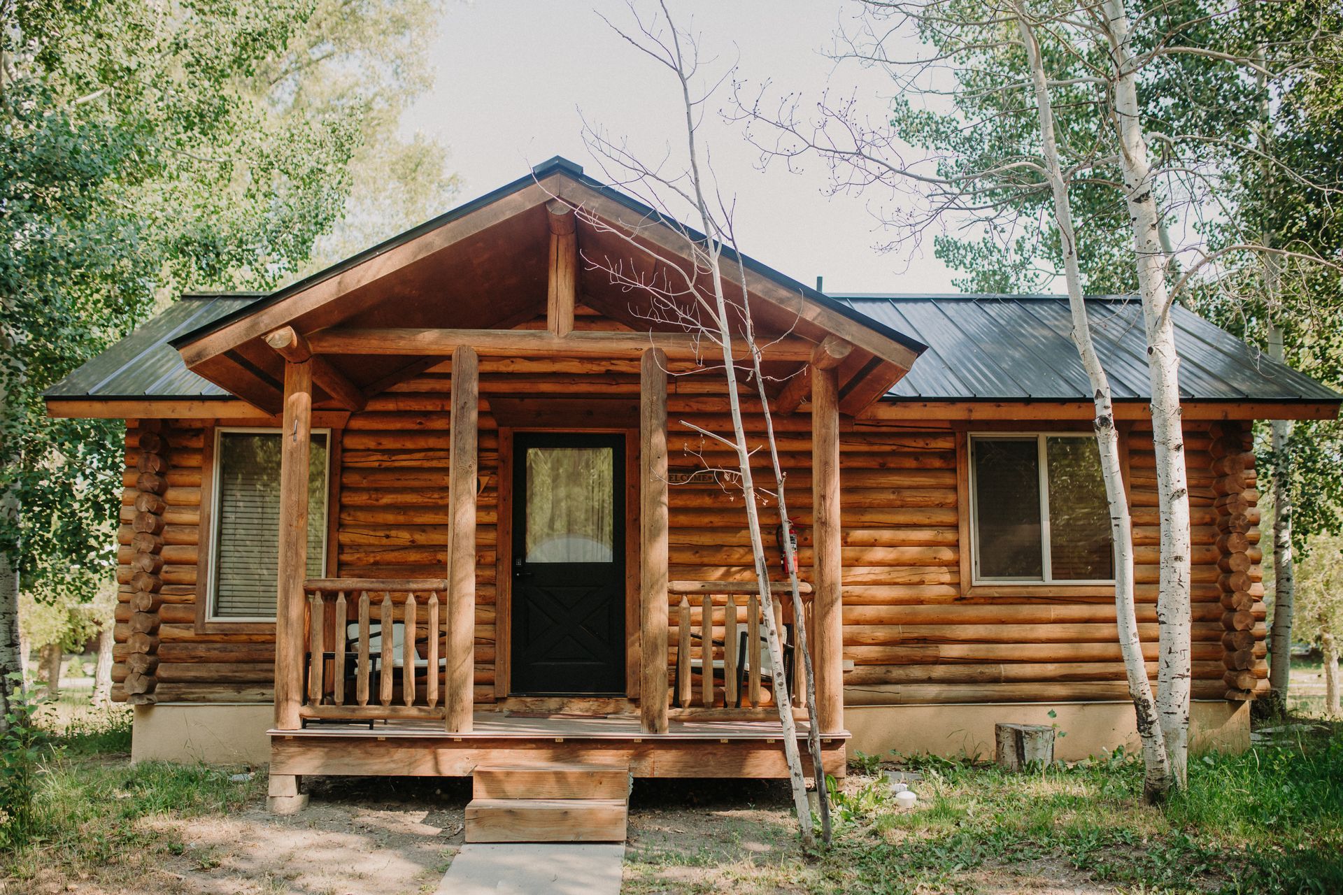 Log cabin with a porch in a wooded setting. Brown logs, dark door, windows.