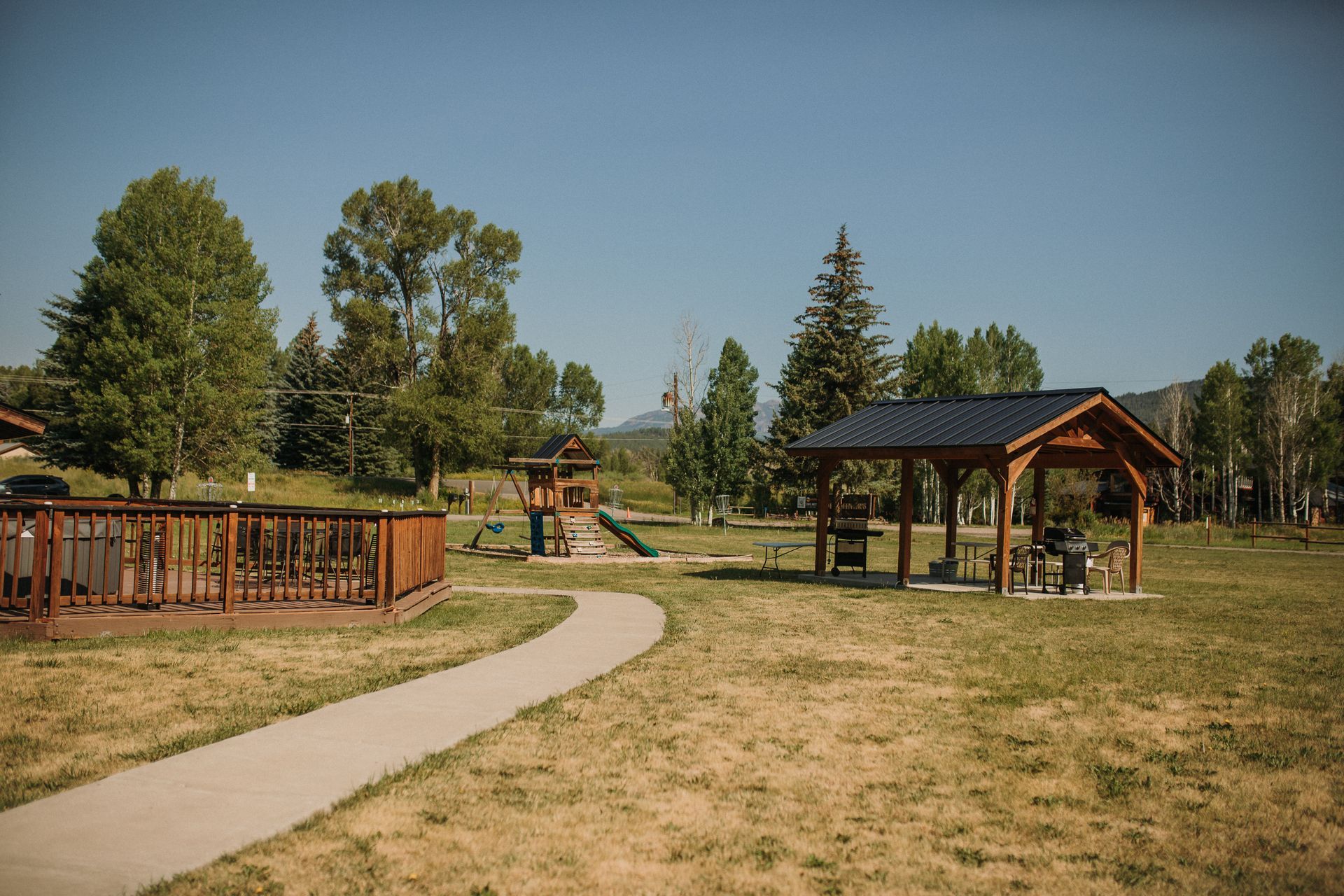 A park with a playground, shelter, and path, under a clear blue sky.