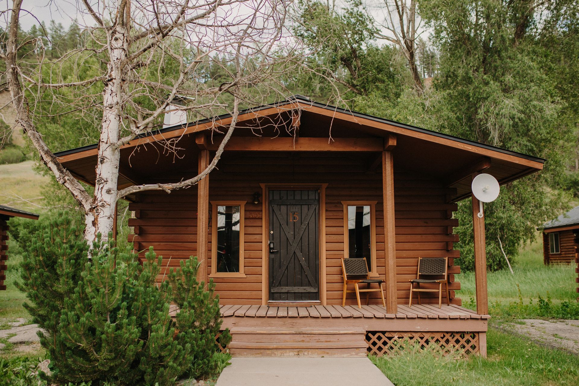 Wooden cabin with porch and two chairs, set in a grassy area with trees and a hill in the background.