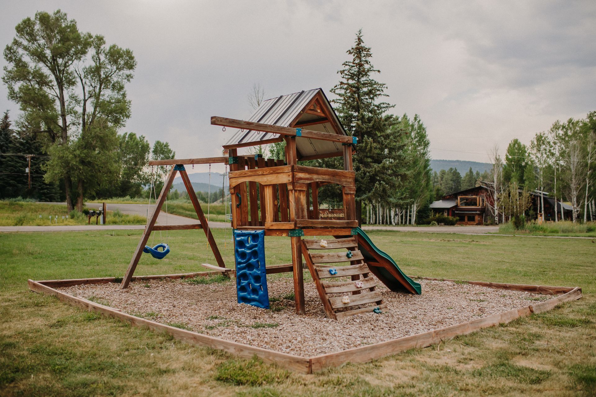 Wooden playground with a swing set, slide, and climbing wall, set on gravel in a grassy field.