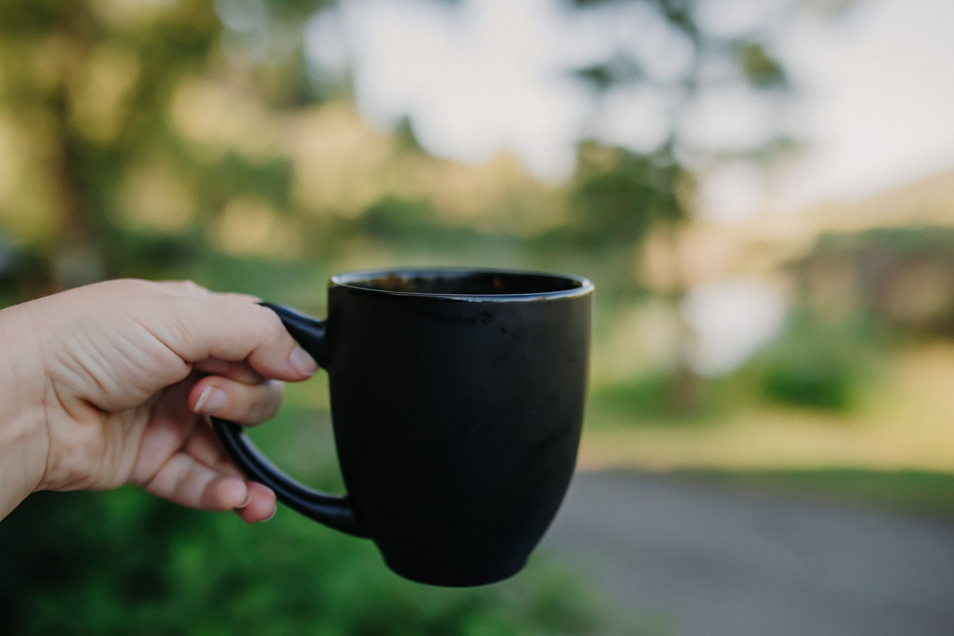 Hand holding a black coffee mug outdoors, overlooking a blurred landscape of trees and water.