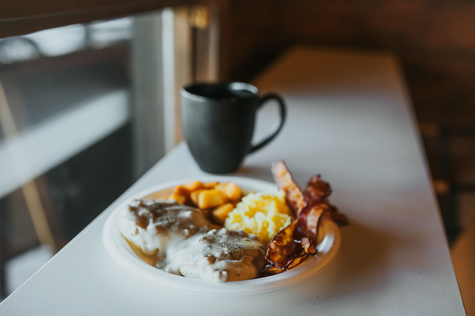 Breakfast plate with gravy, potatoes, bacon, and a black mug of coffee on a white table.