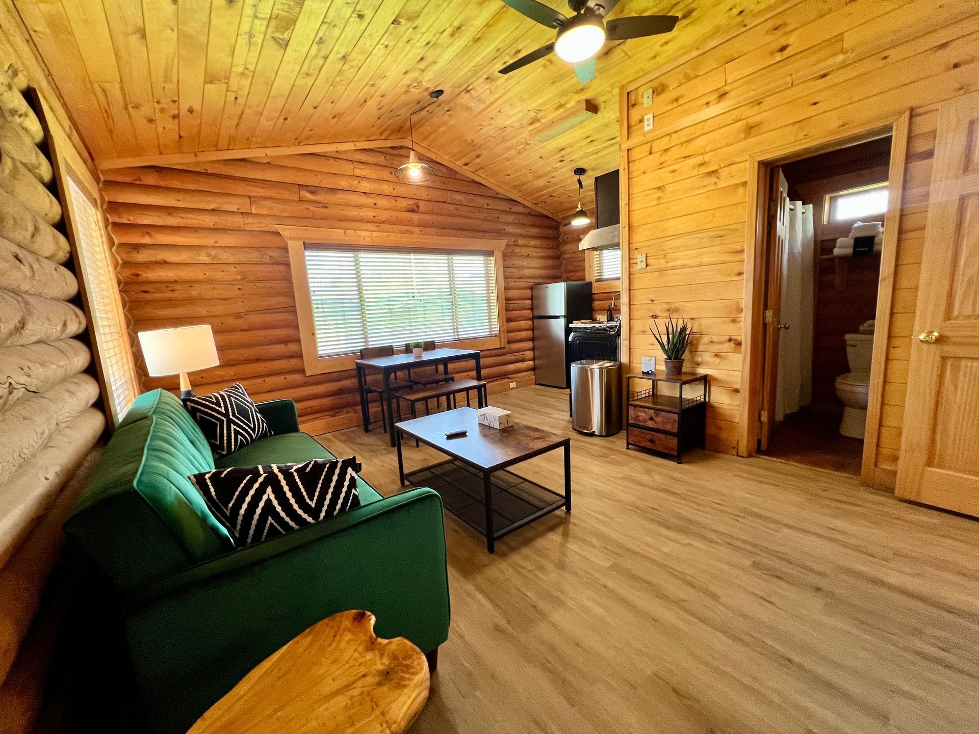 Cozy cabin living room: green sofa, wood walls, ceiling fan, small kitchen, doorway to bathroom.