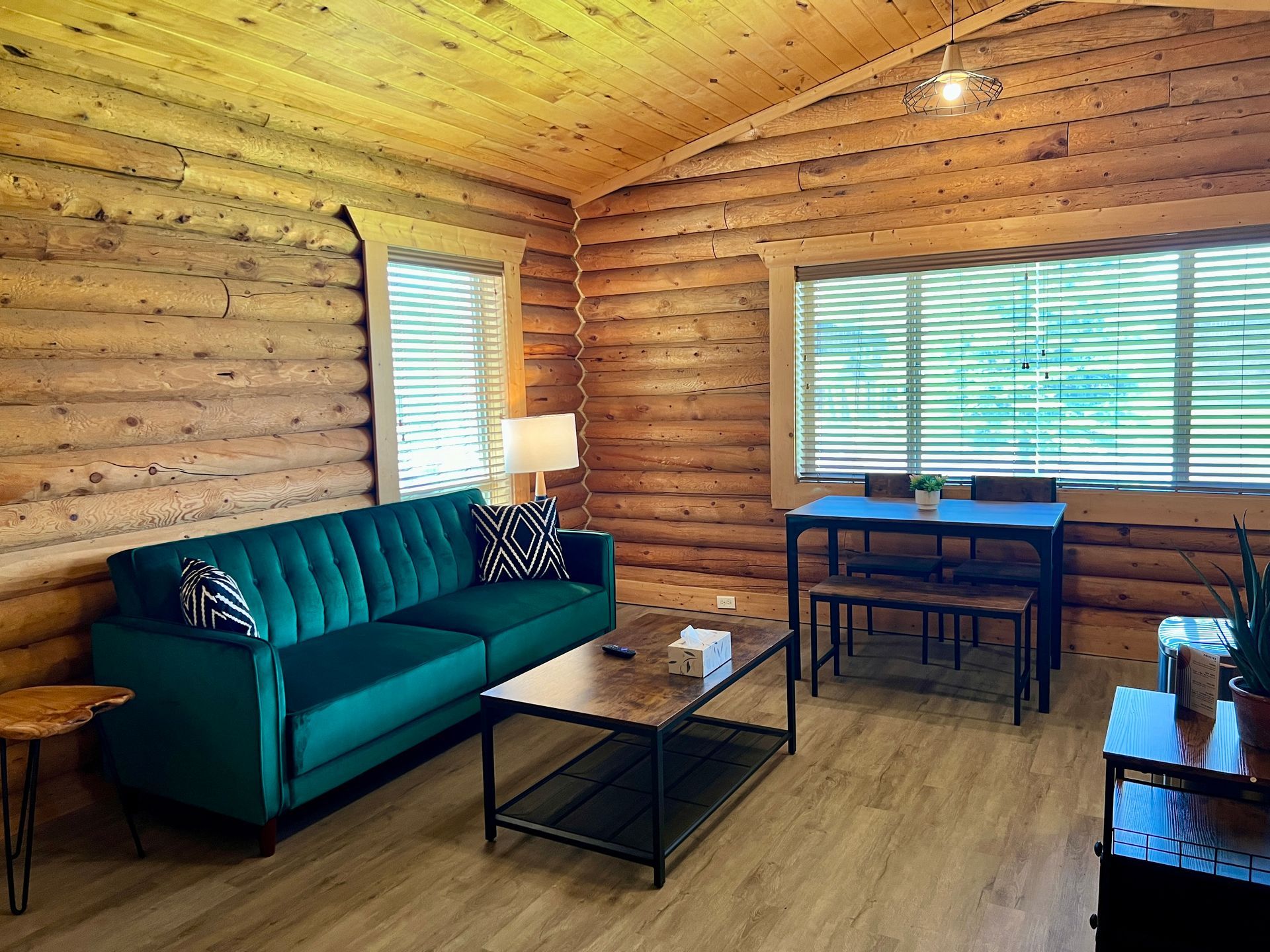 Cozy cabin interior: green sofa, dining table, wood paneling, large windows with blinds, wooden floors.