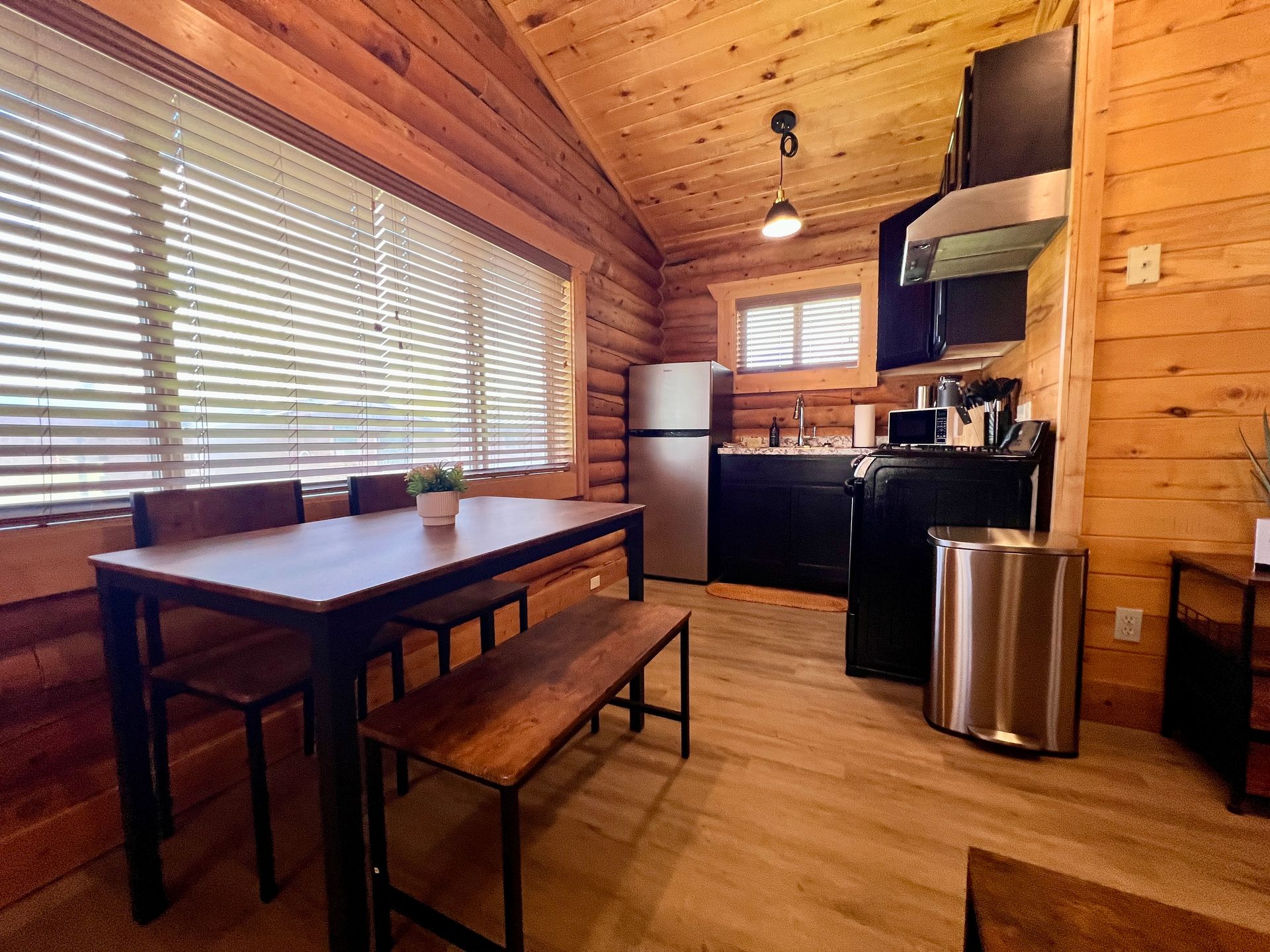 Cozy cabin interior with kitchen, dining area: wooden walls, stainless steel appliances, table and bench.