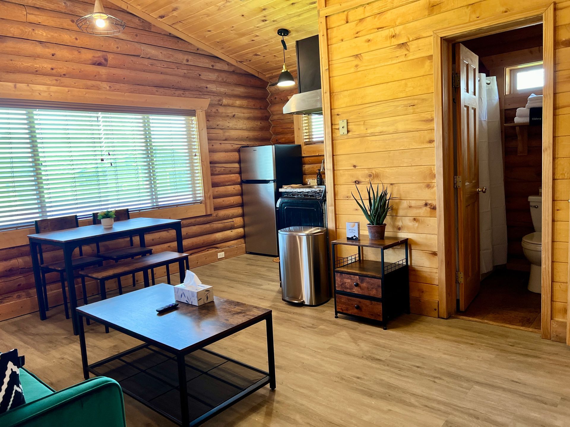 Cozy cabin interior with kitchen, dining area, and open bathroom. Wooden walls and floor, neutral colors.