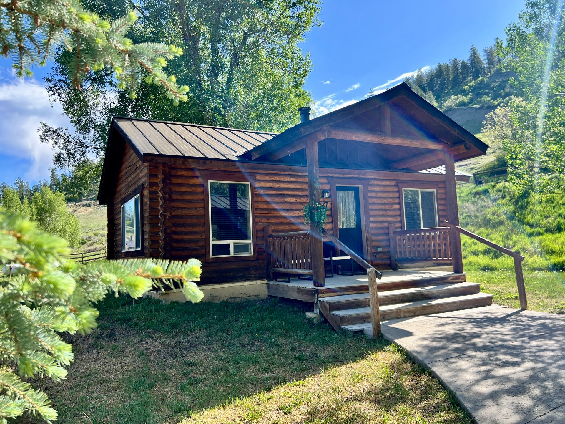 Log cabin with porch in grassy setting, steps up to the door, trees in background.