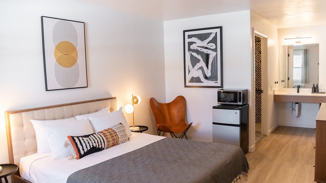 Bedroom with a bed, artwork, a brown chair, and a microwave in a brightly lit setting.