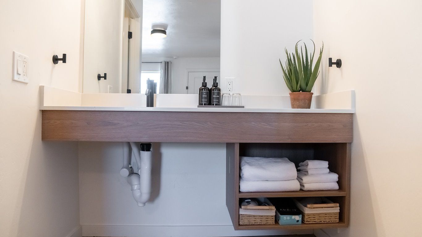 Bathroom vanity with floating wood cabinet, white countertop, mirror, toiletries, towels, and a plant.